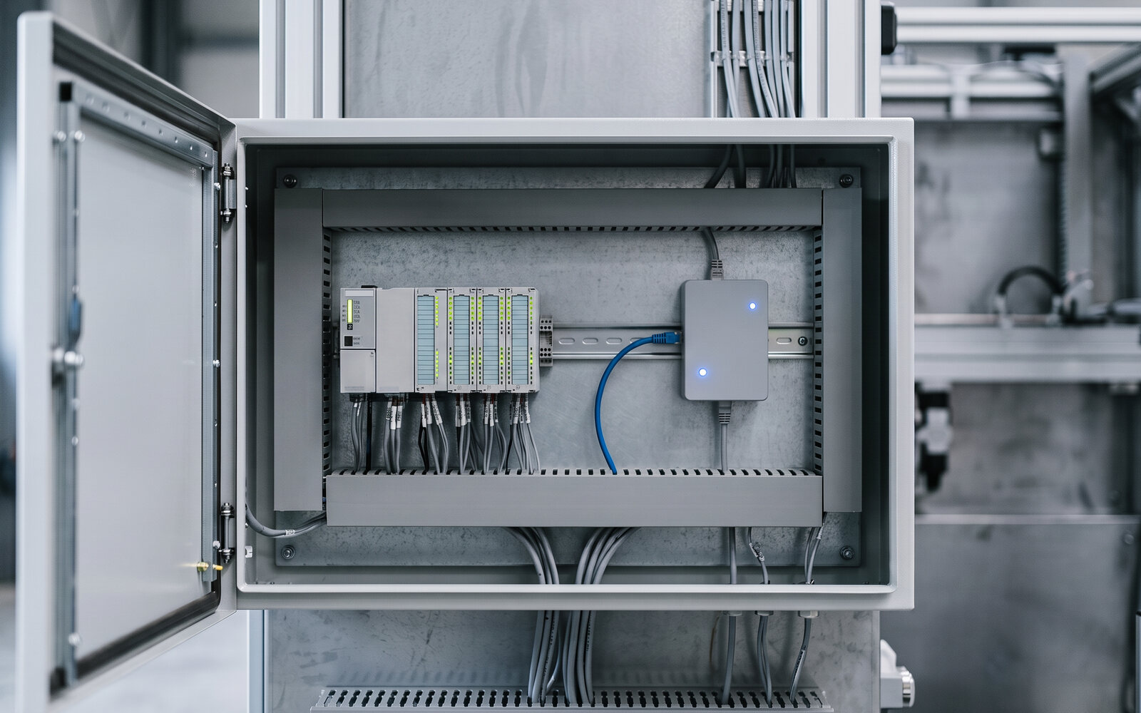 Inside the open control cabinet of a machine: the compact Mavex gateway with its blue status LED, wired with a blue Ethernet cable to the modular PLC on the DIN rail. Illustrative photo.